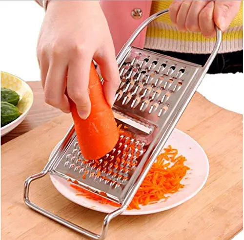 A person is using a Techmanistan 3in1 Stainless Steel Vegetable Slicer Chopper Grater to shred a carrot over a white plate on a wooden cutting board. The shredded carrot collects on the plate, and cucumbers are visible on another plate in the background. This versatile kitchen tool makes meal prep easy and efficient.