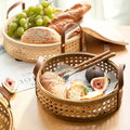 Three woven baskets on a table contain bread, grapes, and figs. One basket has a pair of chopsticks laid across it, highlighting the charm of these Techmanistan Hand Woven Round Rattan Baskets. The setup is accompanied by a partially open book and bright, natural lighting.