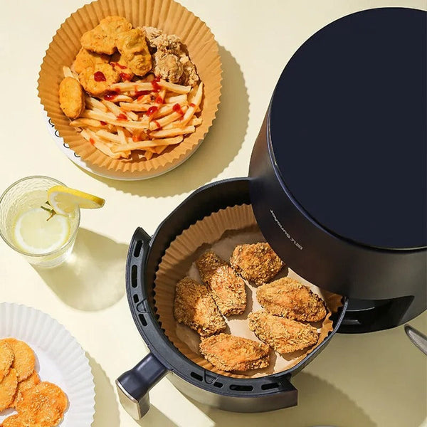 A top-view image of an air fryer with chicken wings inside, placed on a beige tabletop lined with Techmanistan Air Fryer Disposable Paper Liner and Oil Sprayer Bottle. Surrounding the air fryer are plates with various foods, including fries and chicken nuggets, and a glass of lemon water. The scene is bright and minimalist.