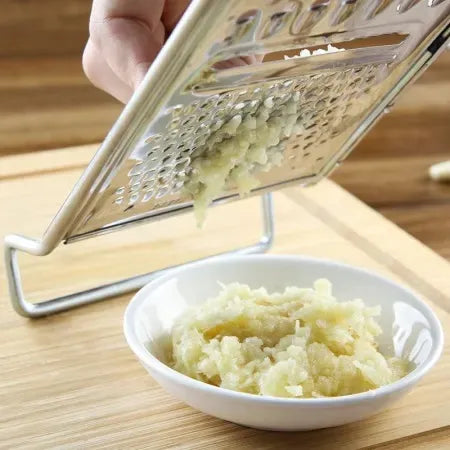 A hand is grating garlic over a small white bowl using the Techmanistan 3in1 Stainless Steel Vegetable Slicer Chopper Grater. Grated garlic is falling into the bowl, making it an essential meal prep accessory. The scene is set on a light wooden surface.