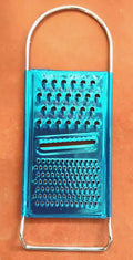 A three-sided blue stainless steel grater with a handle on an orange surface. This versatile kitchen tool features three different grating surfaces, including large holes, small holes, and a slicing slot in the middle, making it the perfect **Techmanistan 3in1 Stainless Steel Vegetable Slicer Chopper Grater**.