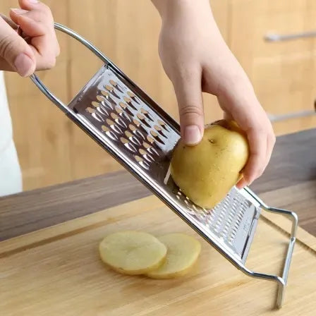 A person is grating a potato with a Techmanistan 3in1 Stainless Steel Vegetable Slicer Chopper Grater over a wooden cutting board. Two slices of potato lie on the board. The background includes a wooden surface and kitchen drawers, showcasing the practicality of this essential kitchen tool.