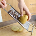 A person is grating a potato with a Techmanistan 3in1 Stainless Steel Vegetable Slicer Chopper Grater over a wooden cutting board. Two slices of potato lie on the board. The background includes a wooden surface and kitchen drawers, showcasing the practicality of this essential kitchen tool.