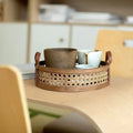 A Hand Woven Round Rattan Basket from Techmanistan, part of a handwoven serving trays set, holds several ceramic cups and a jug on a wooden table. The background includes light-colored wooden furniture and a soft focus on various containers and decor items on shelves.