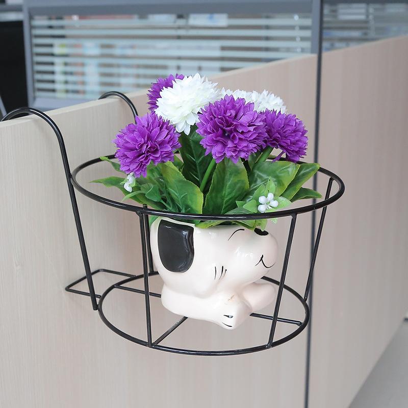 A small, round metal Balcony Railing Fence Flower Pot Stand with an anti-rust coating from My Store hangs on the side of a light-colored wooden surface, holding a ceramic vase shaped like a dog with closed eyes. The vase contains a bouquet of purple and white flowers with green leaves. In the background are horizontal window blinds.