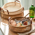 Two stylish Techmanistan Hand Woven Round Rattan Baskets with brown leather handles are placed on a table. One basket contains a bottle of green drink, bread, white cheese, and figs, while the other is empty, nestled underneath. Sunlight streams in, highlighting the cozy setting.