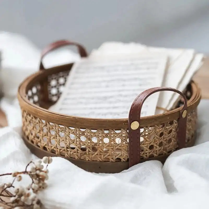 A Hand Woven Round Rattan Basket by Techmanistan with brown leather handles contains an open book with its pages slightly askew. The basket, reminiscent of a handwoven serving tray, is placed on a soft white fabric, and some small dried flowers are visible in the foreground. The background is blurred and neutral.