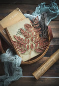 A rustic scene shows a basket containing books, a dried fern leaf, and a speckled feather. An old-fashioned ruler and a piece of blue fabric are placed beside the basket on a wooden surface. The overall aesthetic is vintage and earthy, much like the charm of Techmanistan's Hand Woven Round Rattan Baskets scattered around.