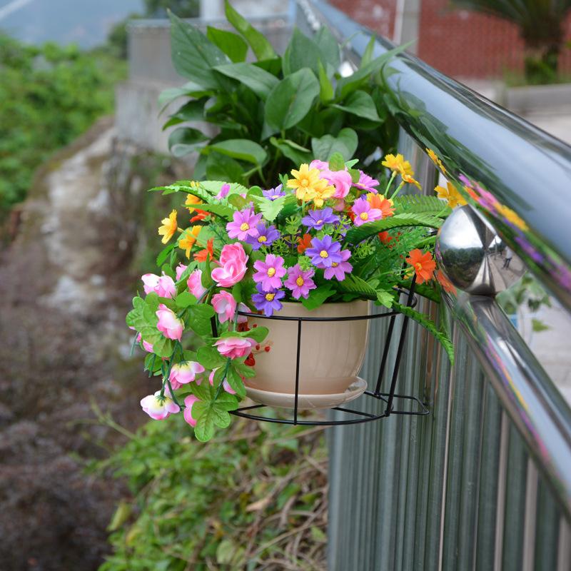 A pot filled with an assortment of colorful flowers, including pink, yellow, and purple blooms, is mounted on a My Store Balcony Railing Fence Flower Pot Stand with a black wire holder. Lush green foliage surrounds the vibrant flowers, while the anti-rust coating ensures durability. The background shows a pathway and greenery.